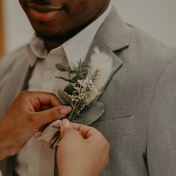 A close-up of a person pinning a boutonniere with white flowers and greenery onto a groom’s light grey suit jacket, symbolising a wedding moment